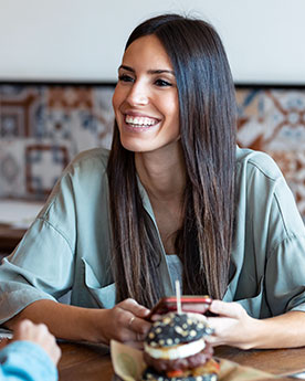 A young lady is smiling at someone while holding her mobile phone and having a meal.